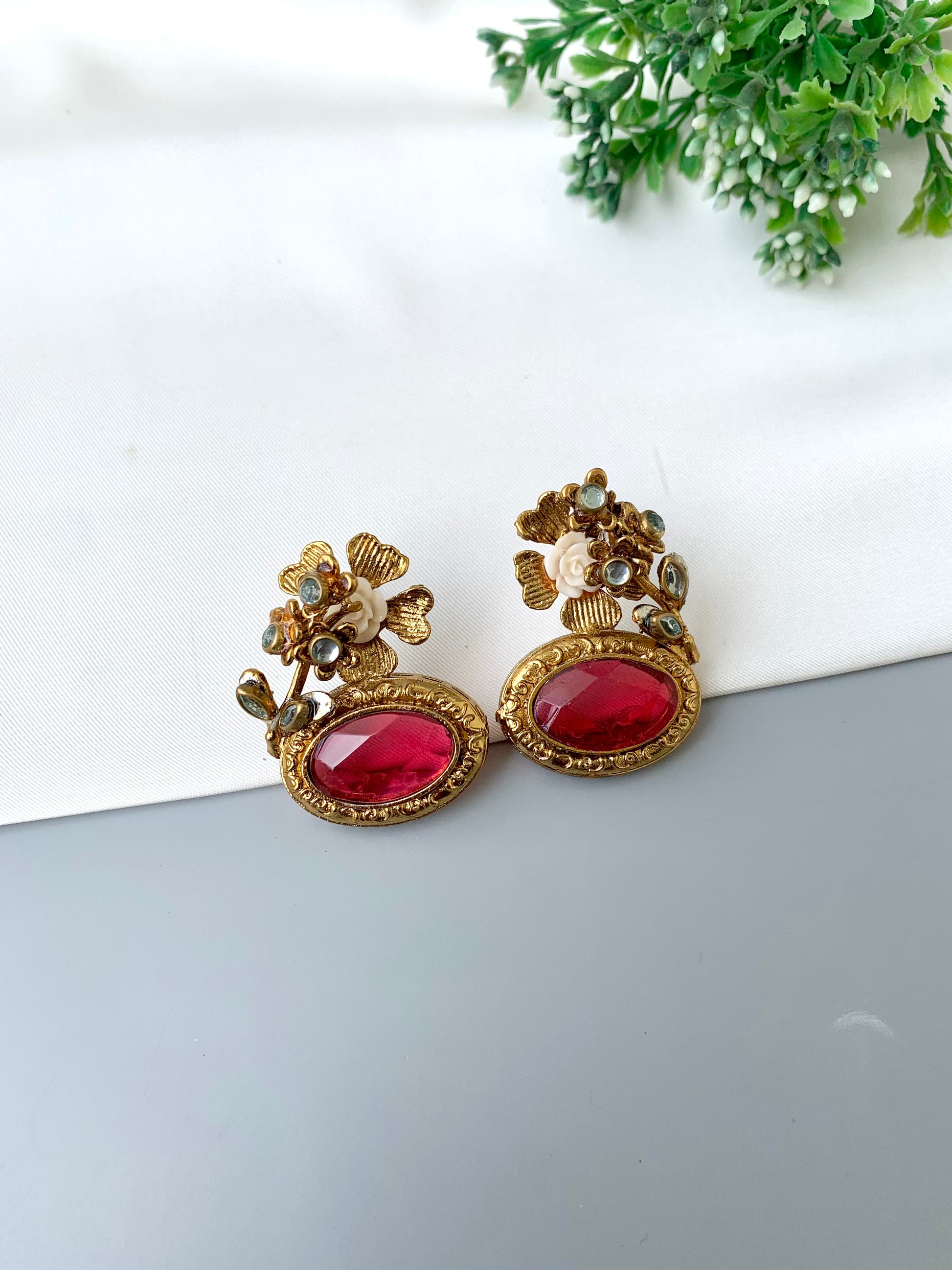 Gold and red earrings on a white surface with greenery in the background