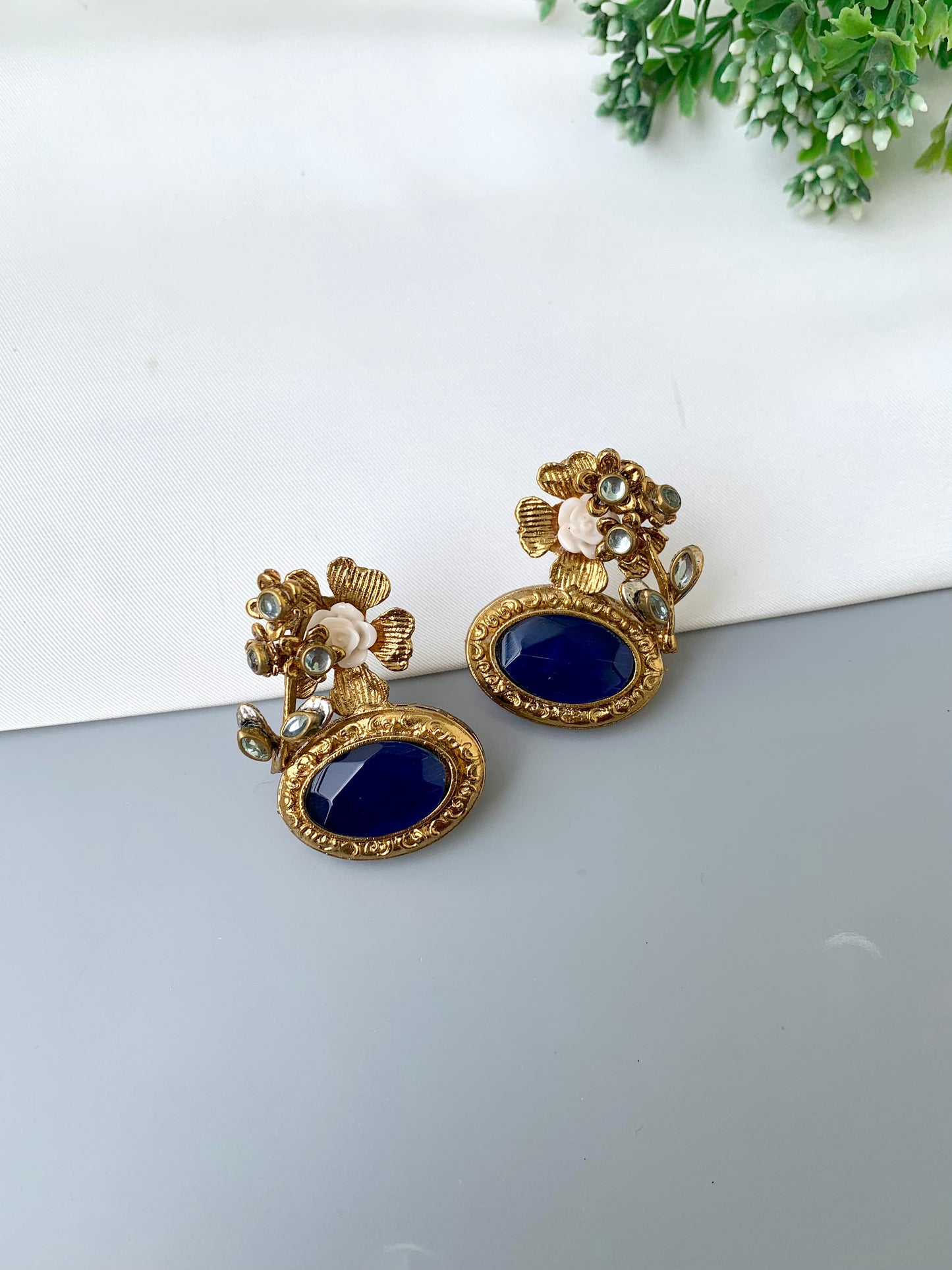 Pair of gold and blue earrings on a white surface with greenery in the corner.