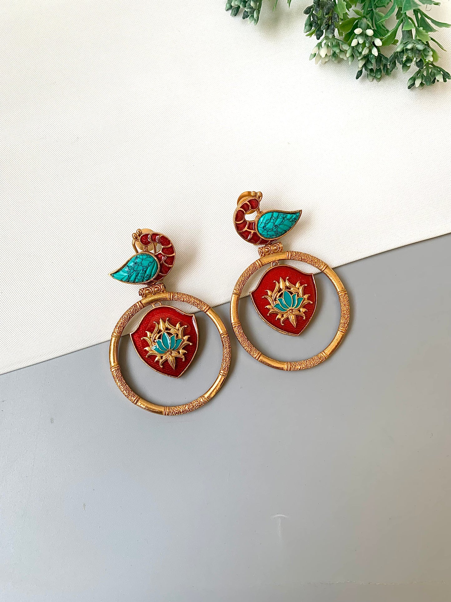Decorative earrings with red and gold design on a white surface with greenery in the background