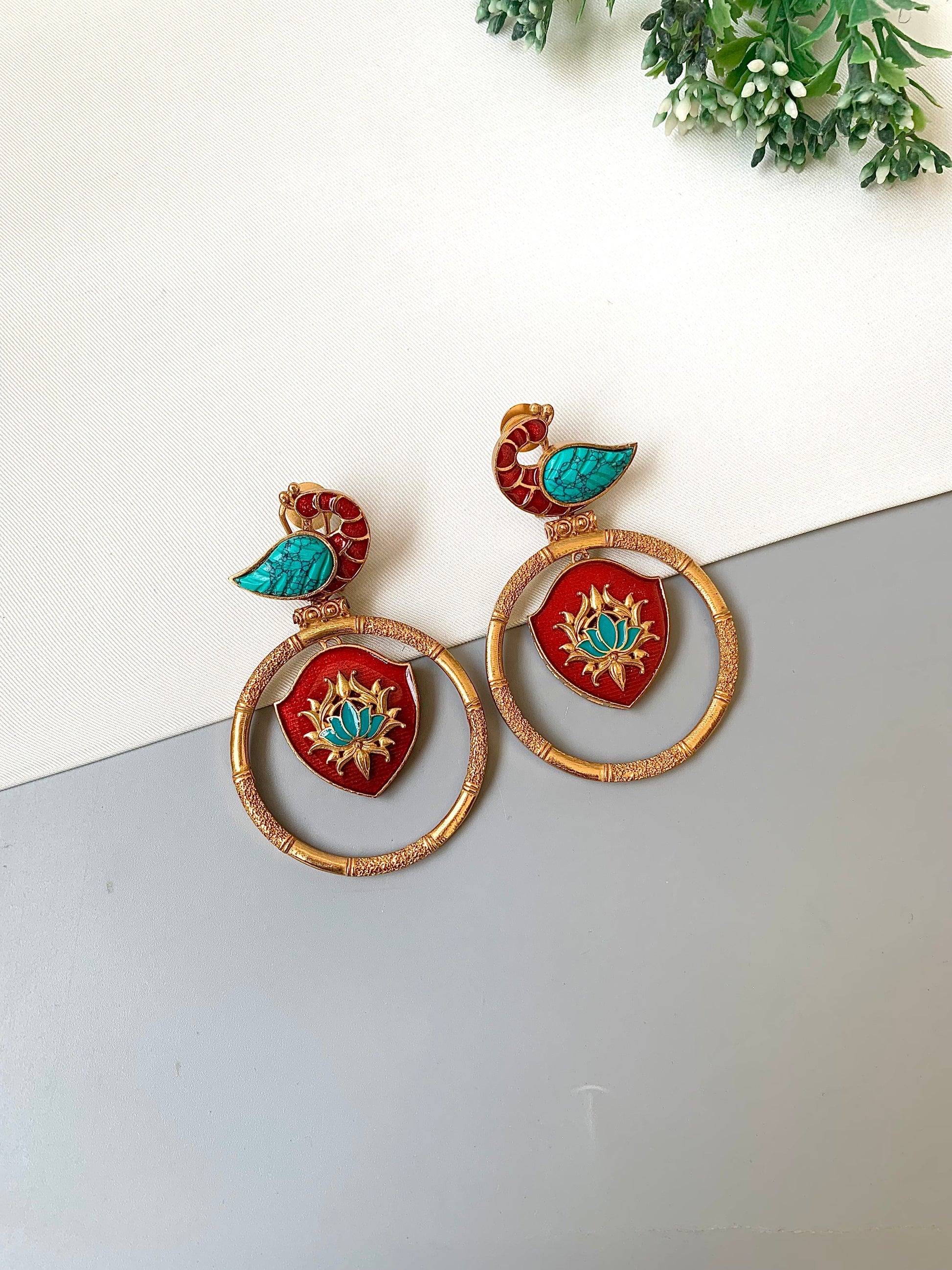Decorative earrings with red and gold design on a white surface with greenery in the background