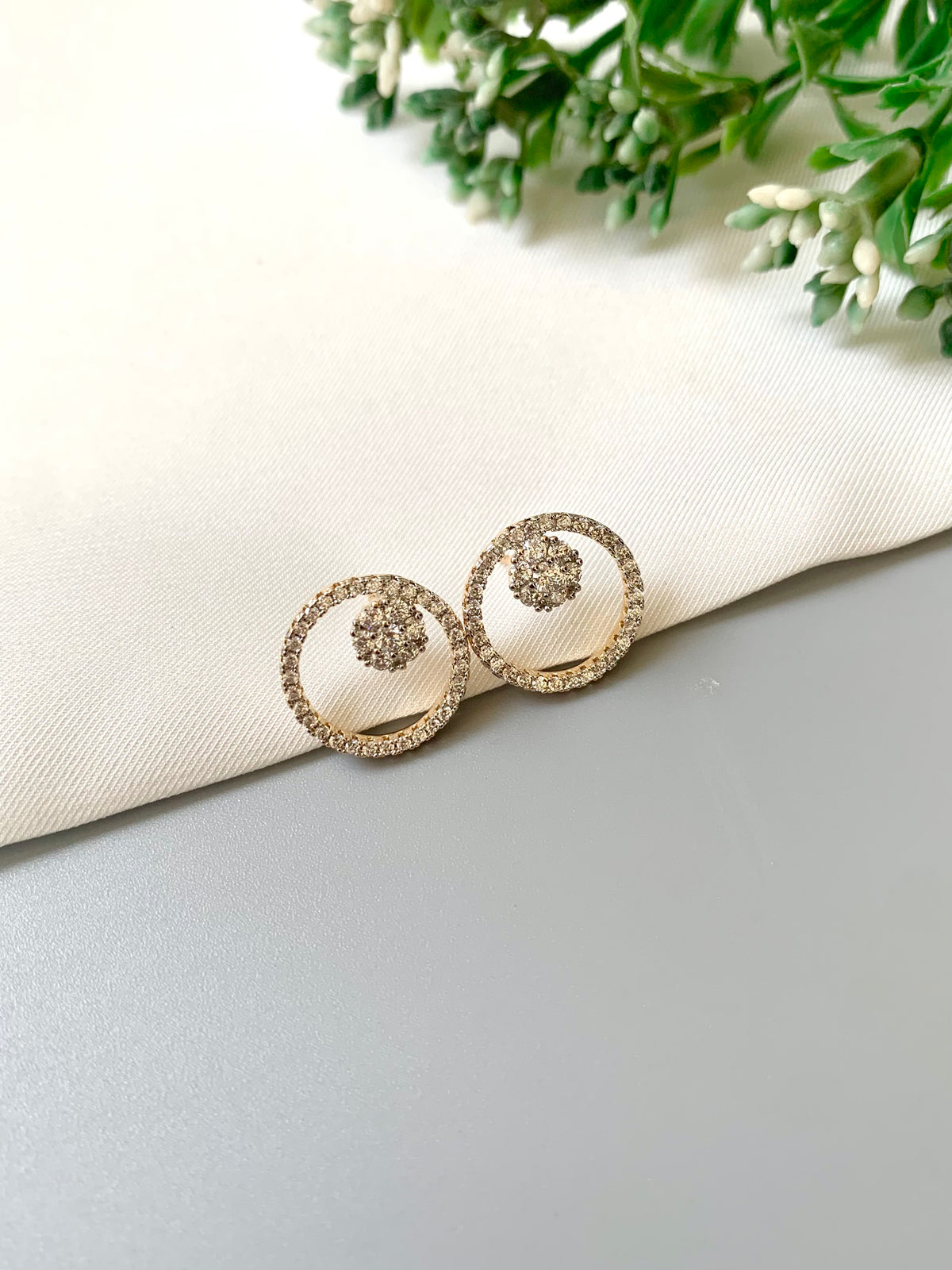 A pair of gold-toned circular stud earrings with small clear stones, resting on a folded cream fabric surface with green foliage in the corner.