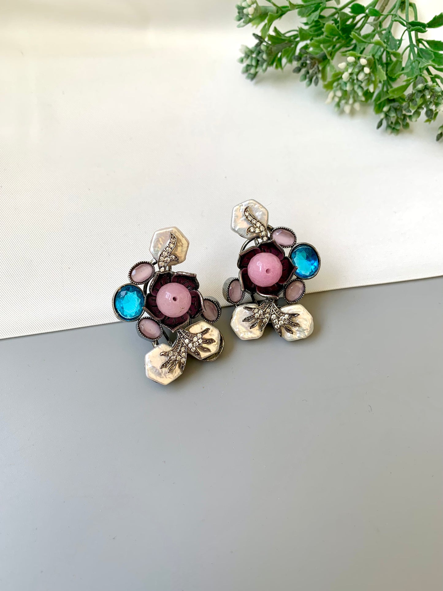 Pair of floral earrings with colorful stones on a white surface with greenery.