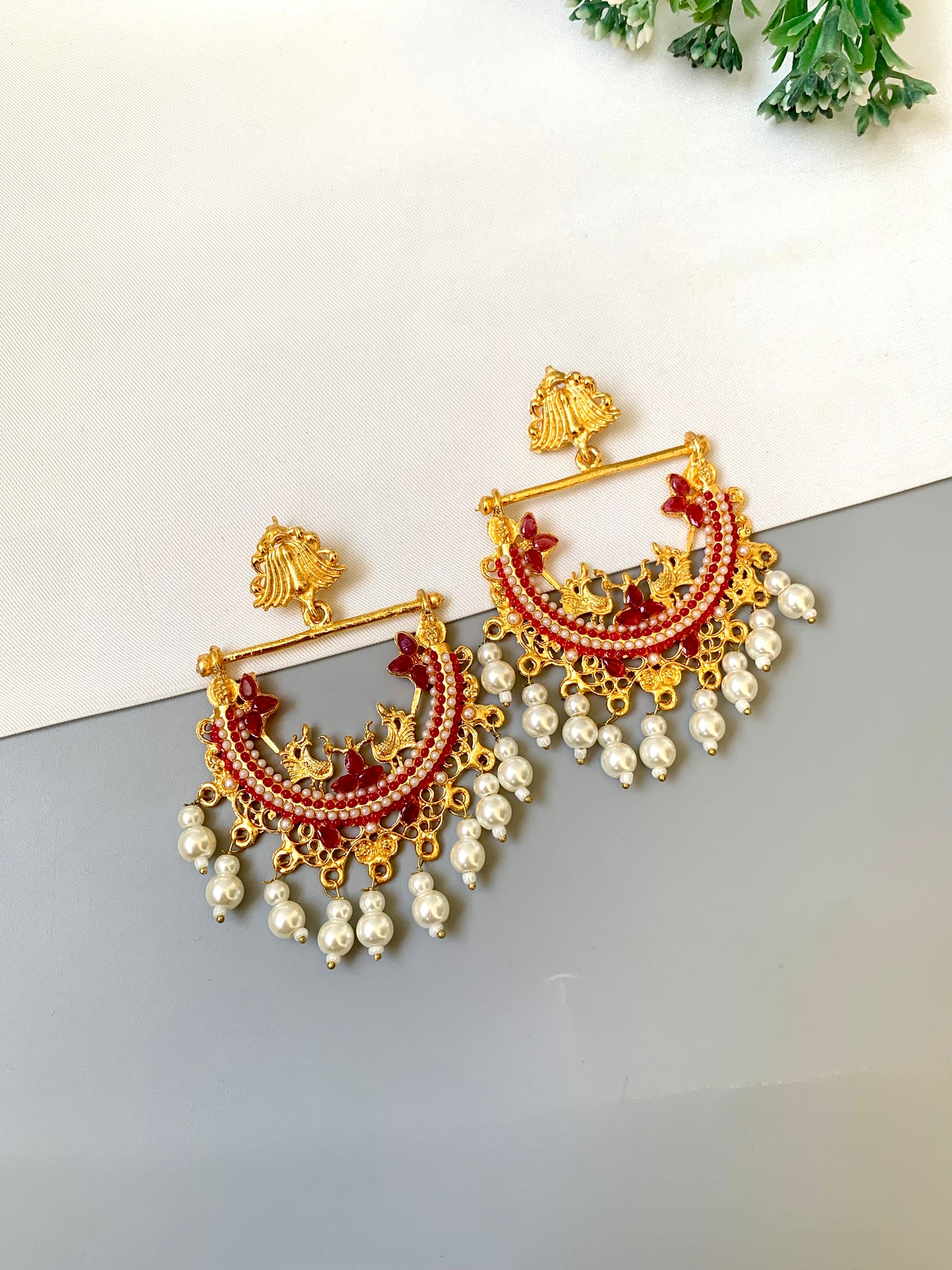 Ornate gold, red, and pearl earrings on a reflective surface with a white background.
