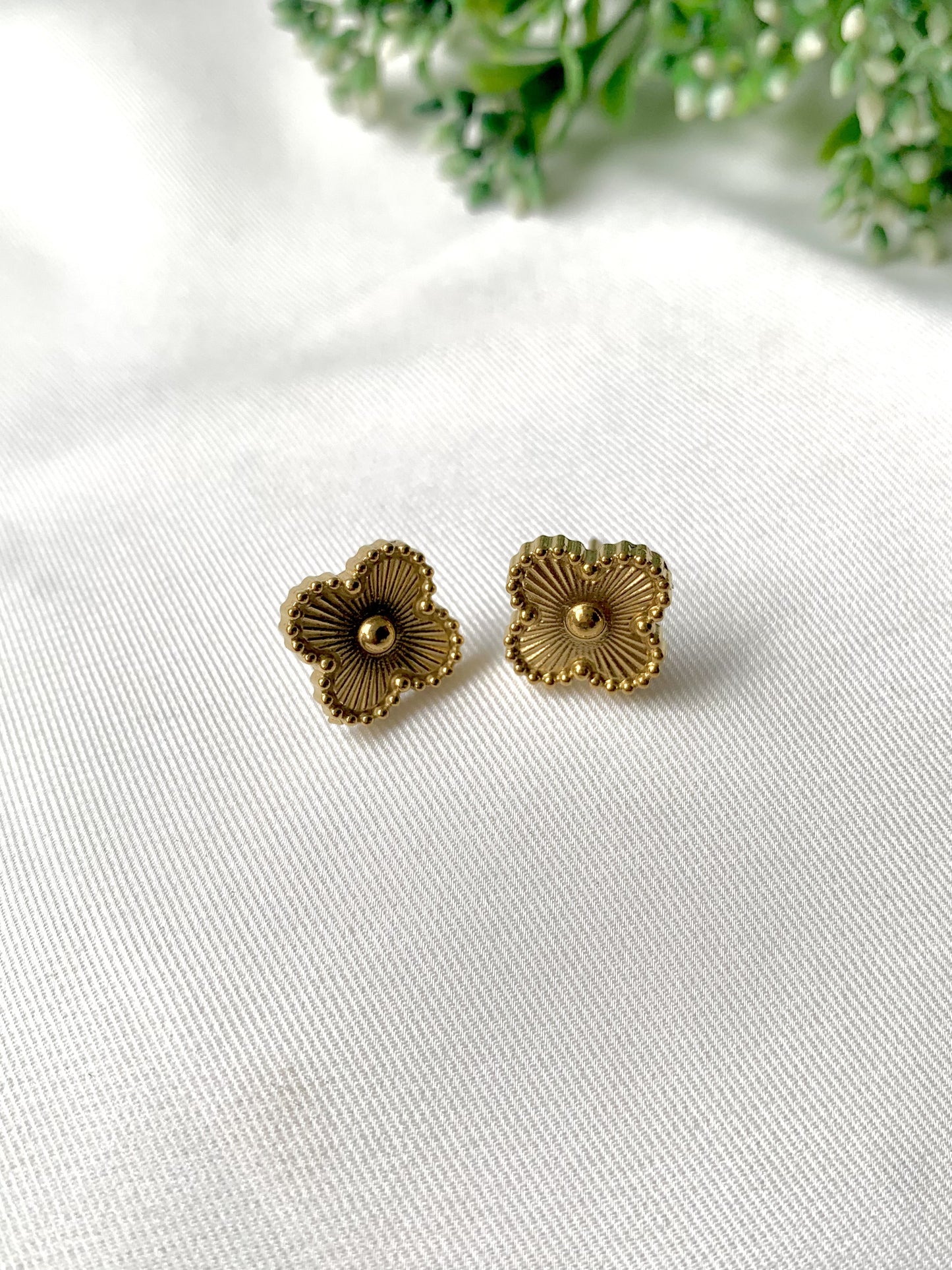 Pair of gold floral earrings on a white fabric background with greenery.