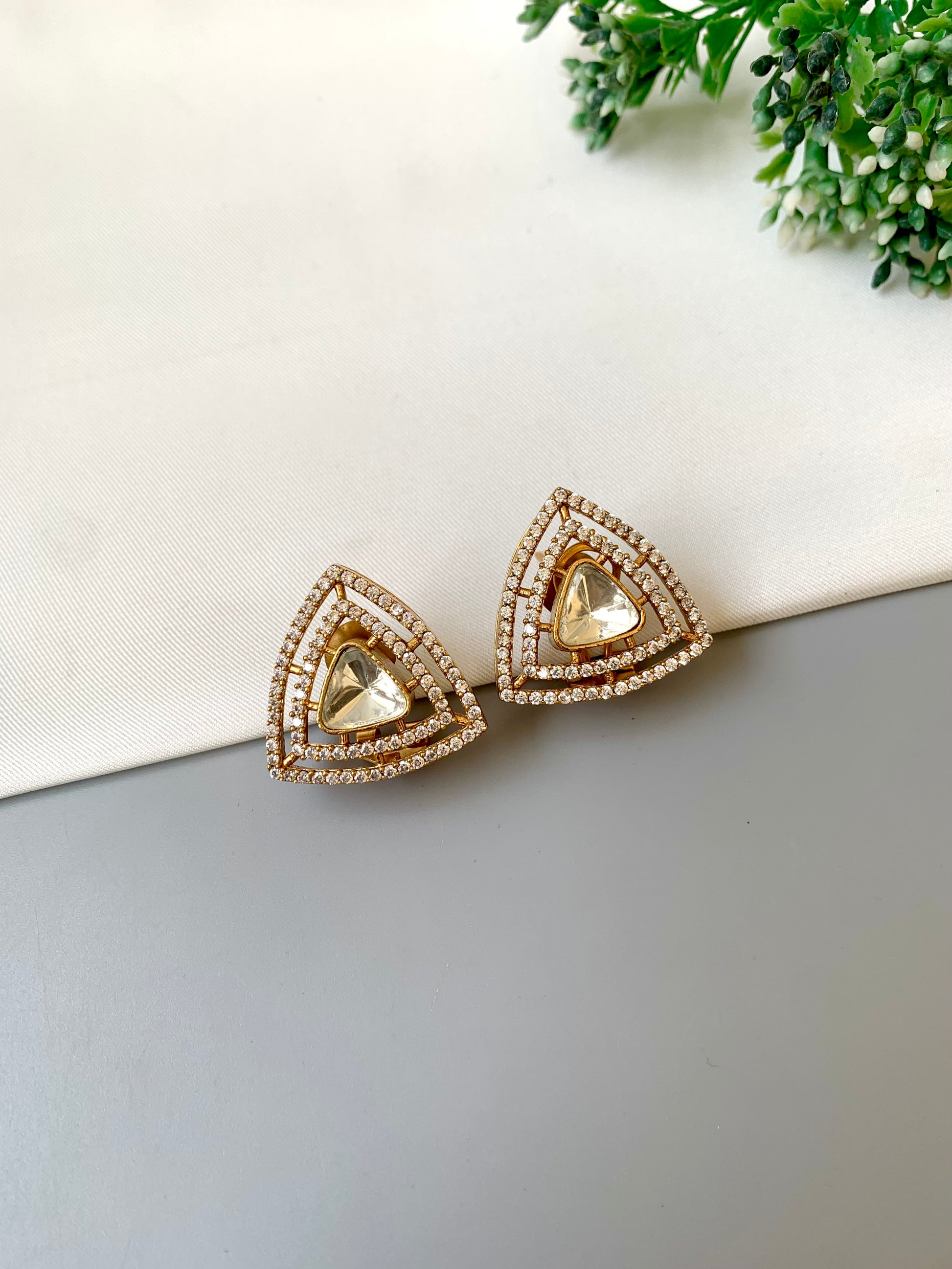 Triangular gold earrings on a white surface with greenery in the background