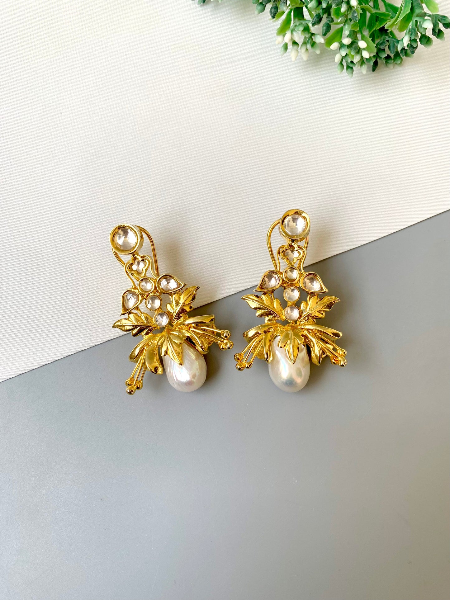 Gold leaf earrings with pearls on a white surface with greenery in the corner.