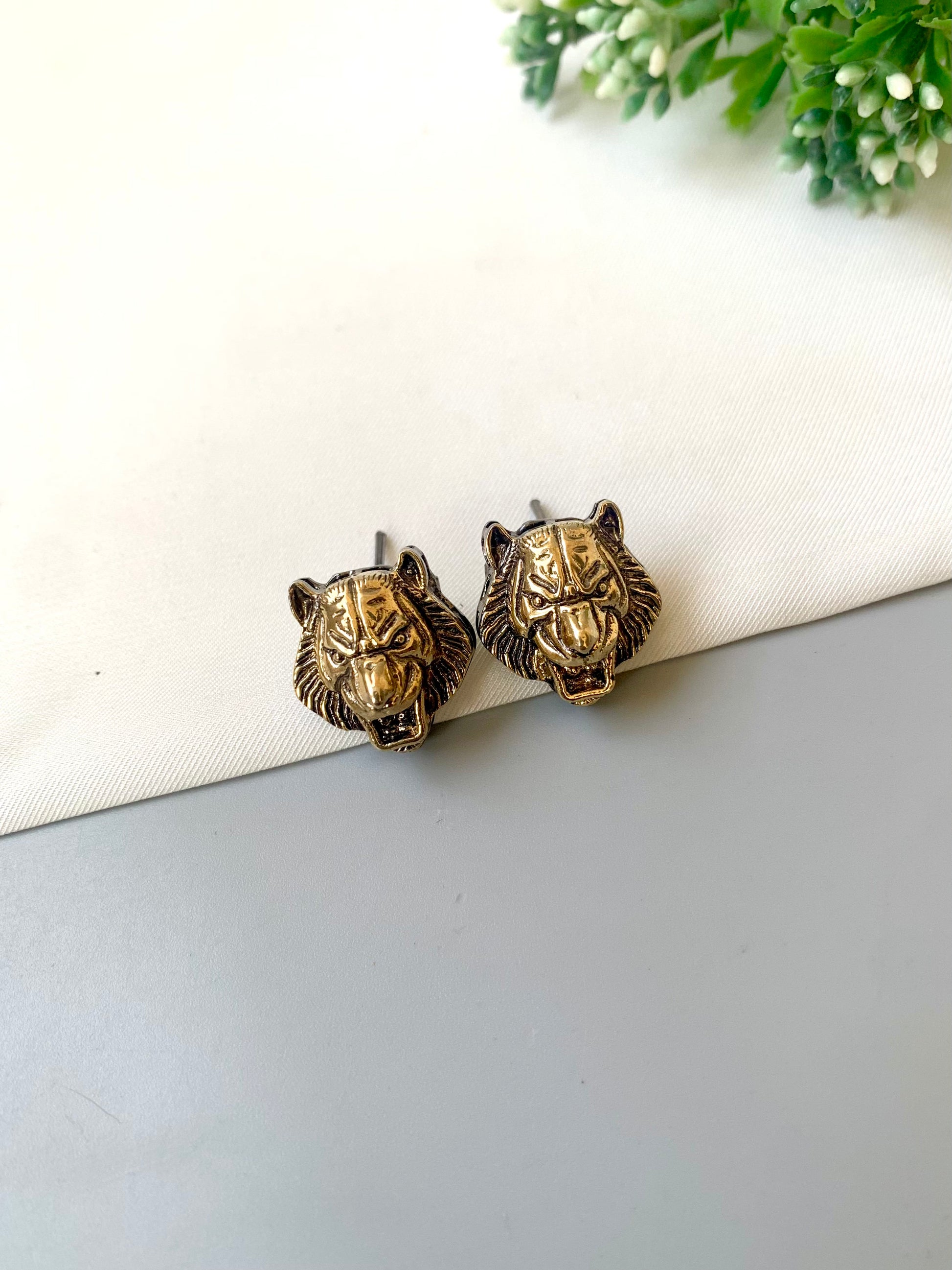Pair of gold lion head earrings on a white surface with greenery in the background