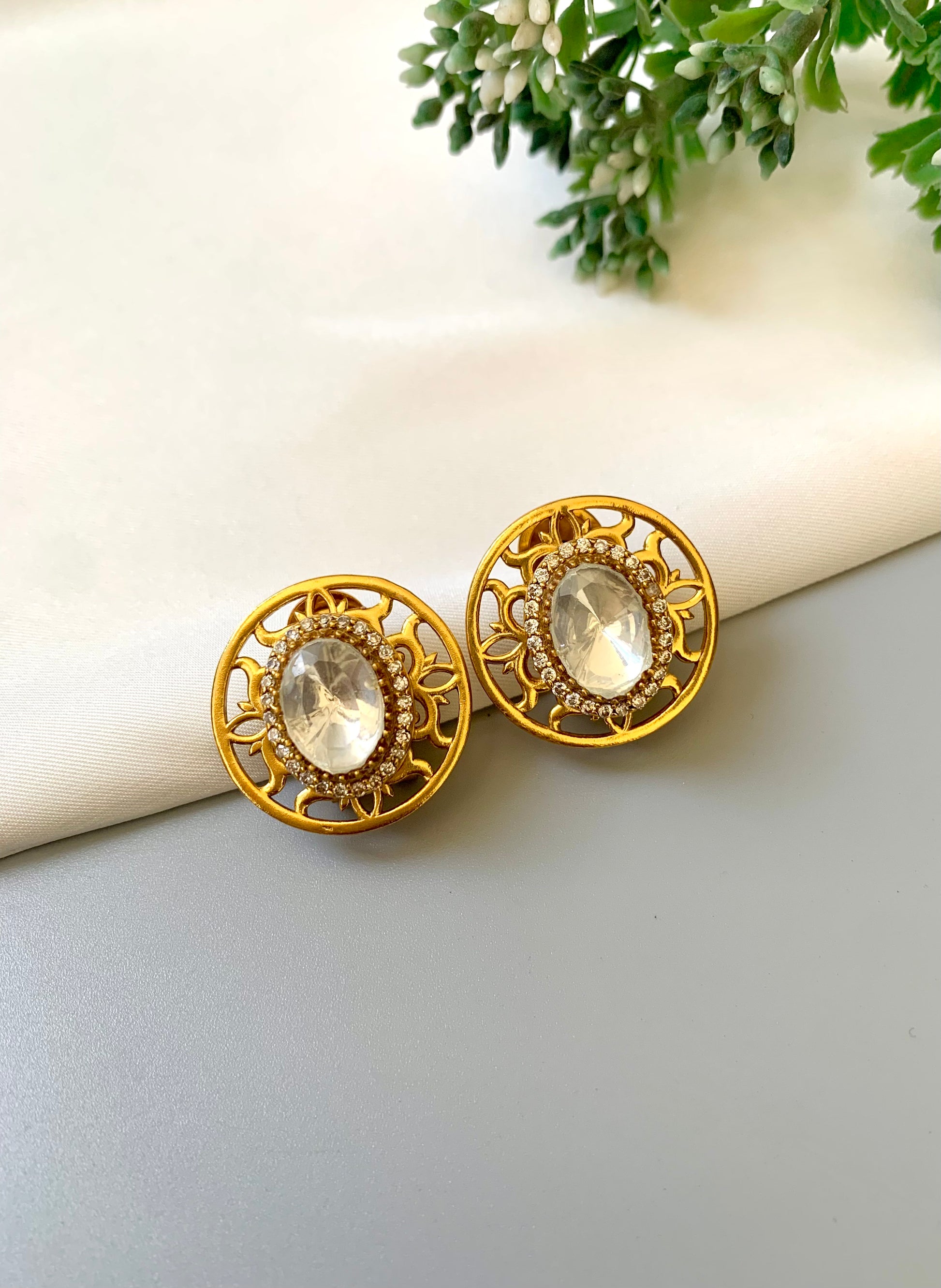 Gold earrings with clear gemstones on a light surface with greenery in the background