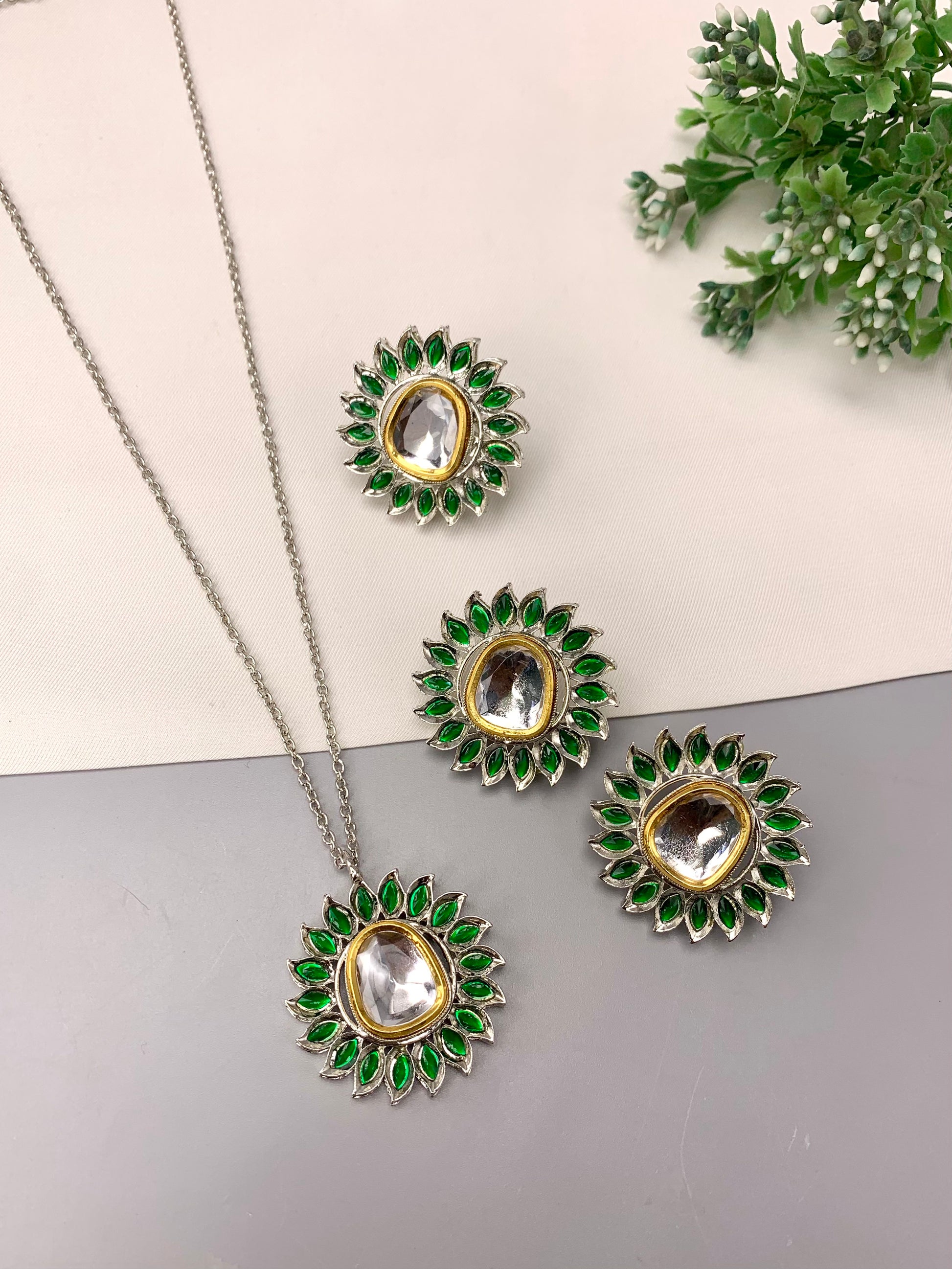 A jewelry set featuring a necklace and three matching earrings with green stone petal details and clear central stones, arranged on a split cream and gray background with a small green plant in the corner.