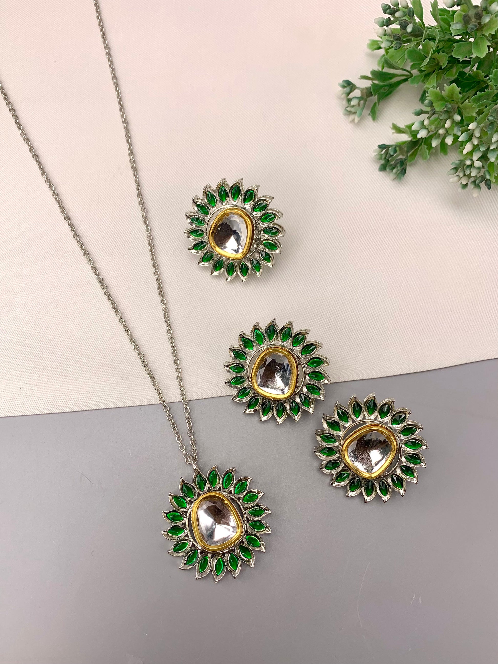 A jewelry set featuring a silver-tone chain necklace, pendant, and two matching stud earrings with green stone accents and clear central stones, arranged on a split beige and gray background with a small green plant in the corner.