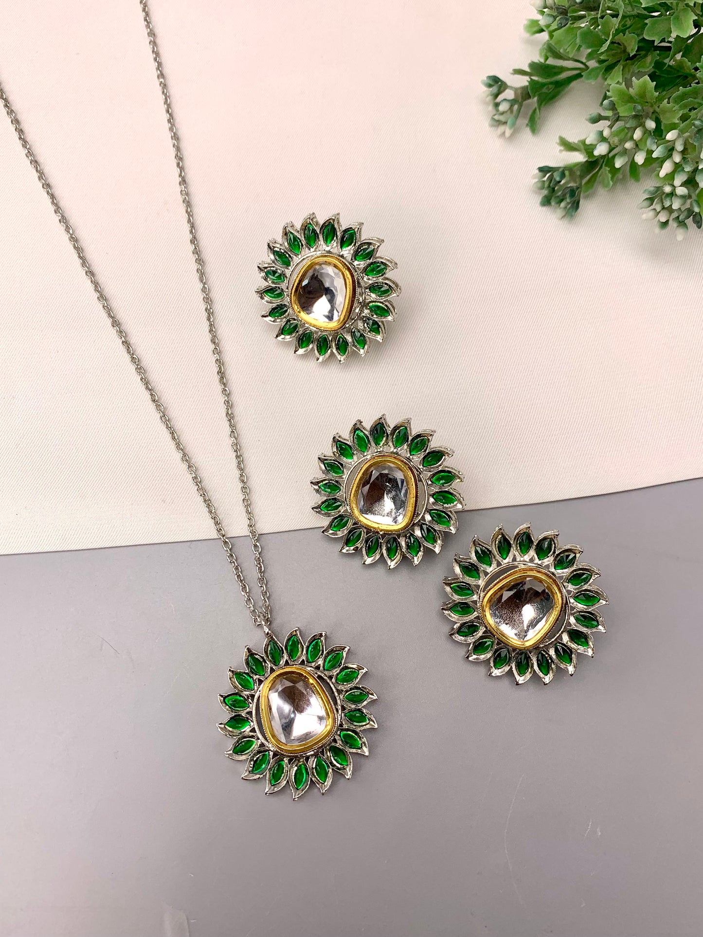 A jewelry set featuring a necklace and three matching earrings with green stone accents and clear central stones, arranged on a split cream and gray background with a small green plant in the corner.