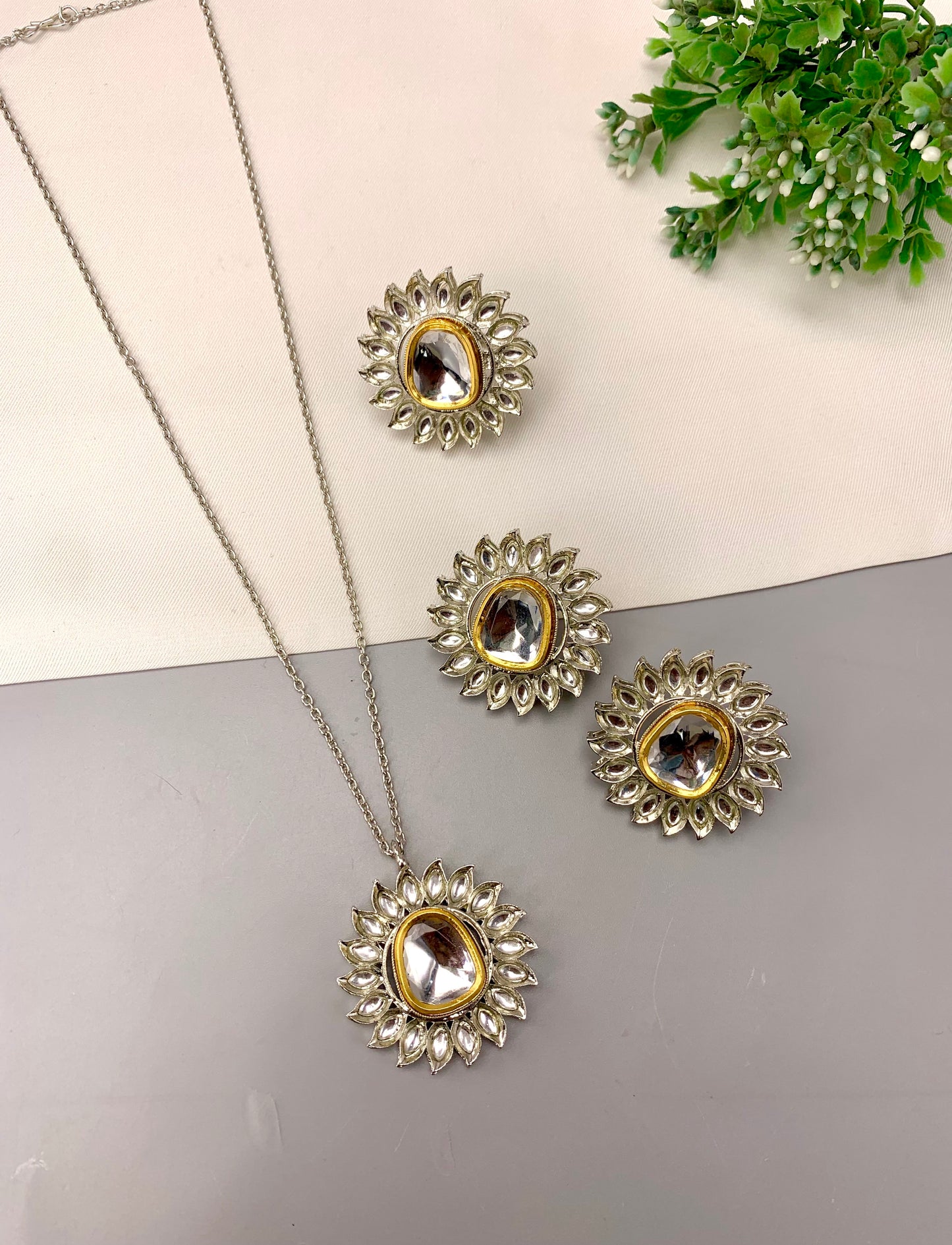 A jewelry set featuring a silver-toned necklace with a sunburst pendant, matching stud earrings, and a small ring, all adorned with clear rhinestones and a central oval stone, displayed on a split cream and gray background with green foliage in the corner.