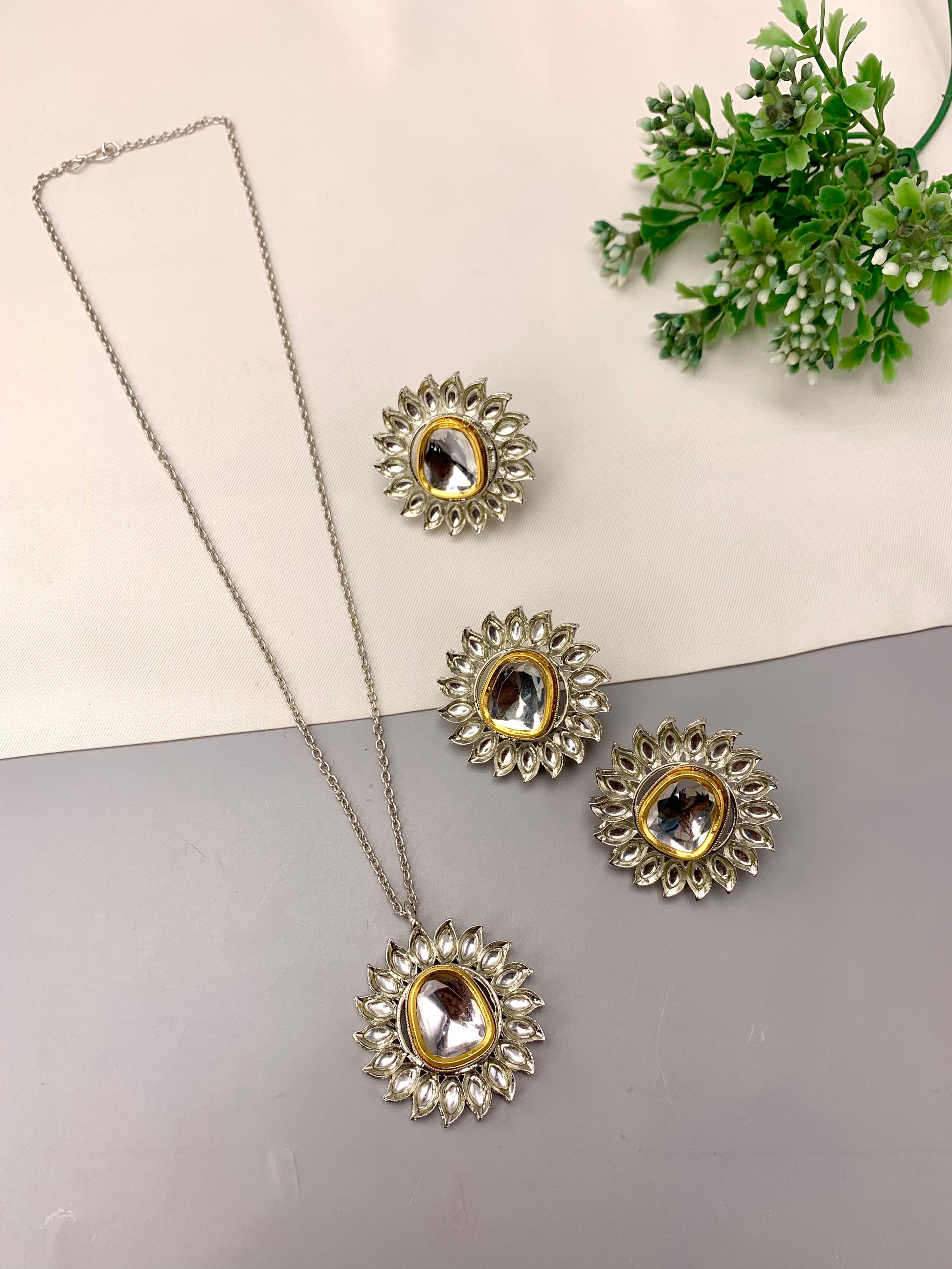 A silver-toned jewelry set featuring a necklace with a sunburst pendant and matching stud earrings, each with a clear central stone and gold-toned detailing, arranged on a split white and gray surface with a small green plant in the corner.