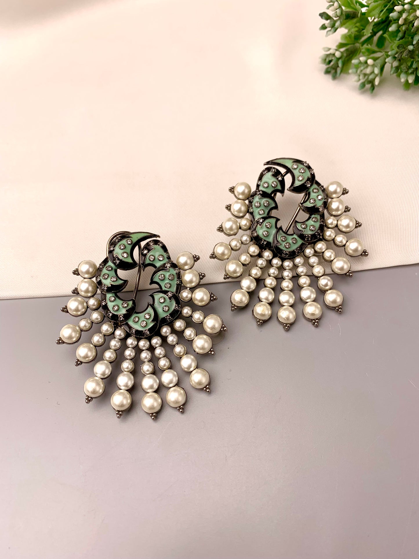 A pair of ornate drop earrings with a black and mint green floral design, embellished with white faux pearls, displayed on a split white and gray surface with a small green plant in the corner.