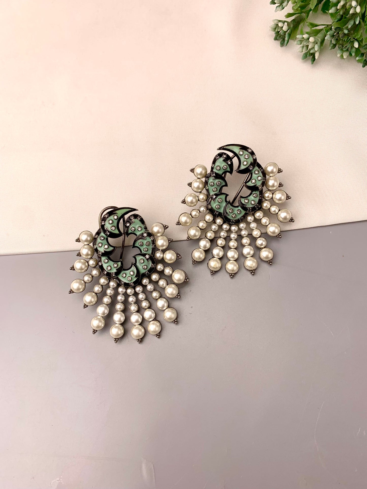 A pair of statement earrings featuring a black filigree top with light green enamel details, adorned with cascading clusters of white faux pearls on a split beige and gray background.
