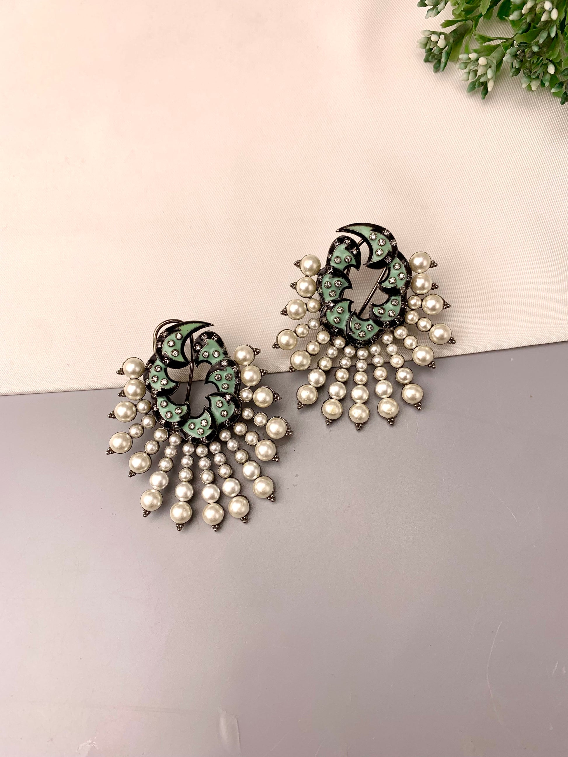 A pair of statement earrings featuring a black filigree top with light green enamel details, adorned with cascading clusters of white faux pearls on a split beige and gray background.