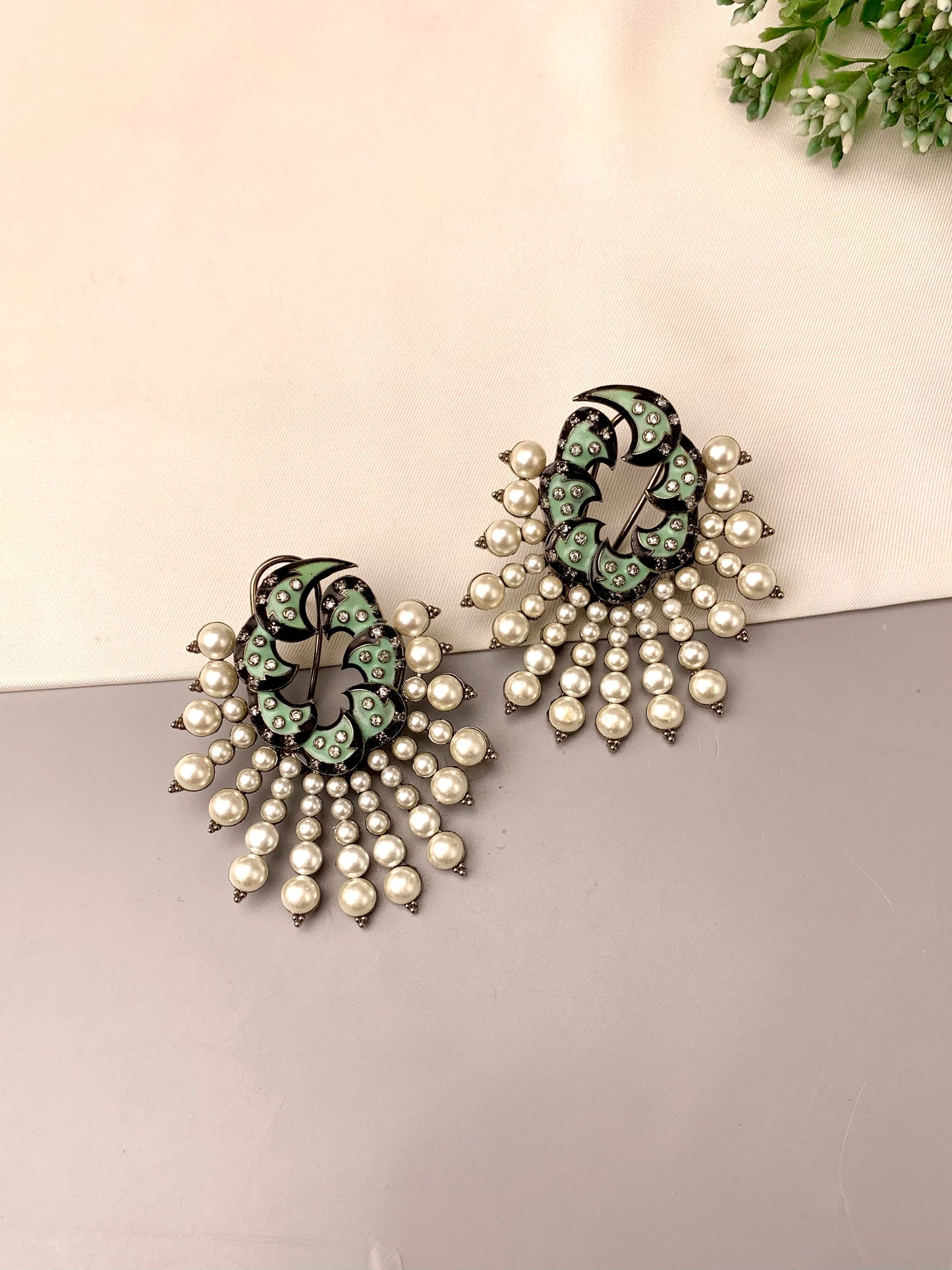 A pair of statement earrings featuring a black and mint green floral enamel design with dangling pearl chains, displayed on a split beige and gray surface.