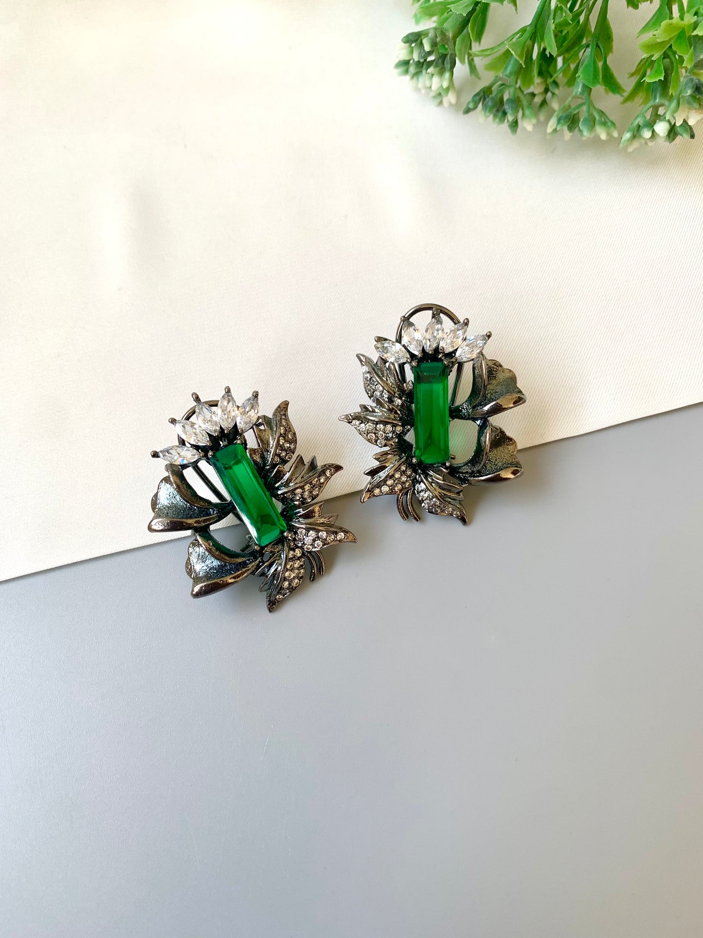 Pair of earrings with green gemstones on a light surface with greenery in the background