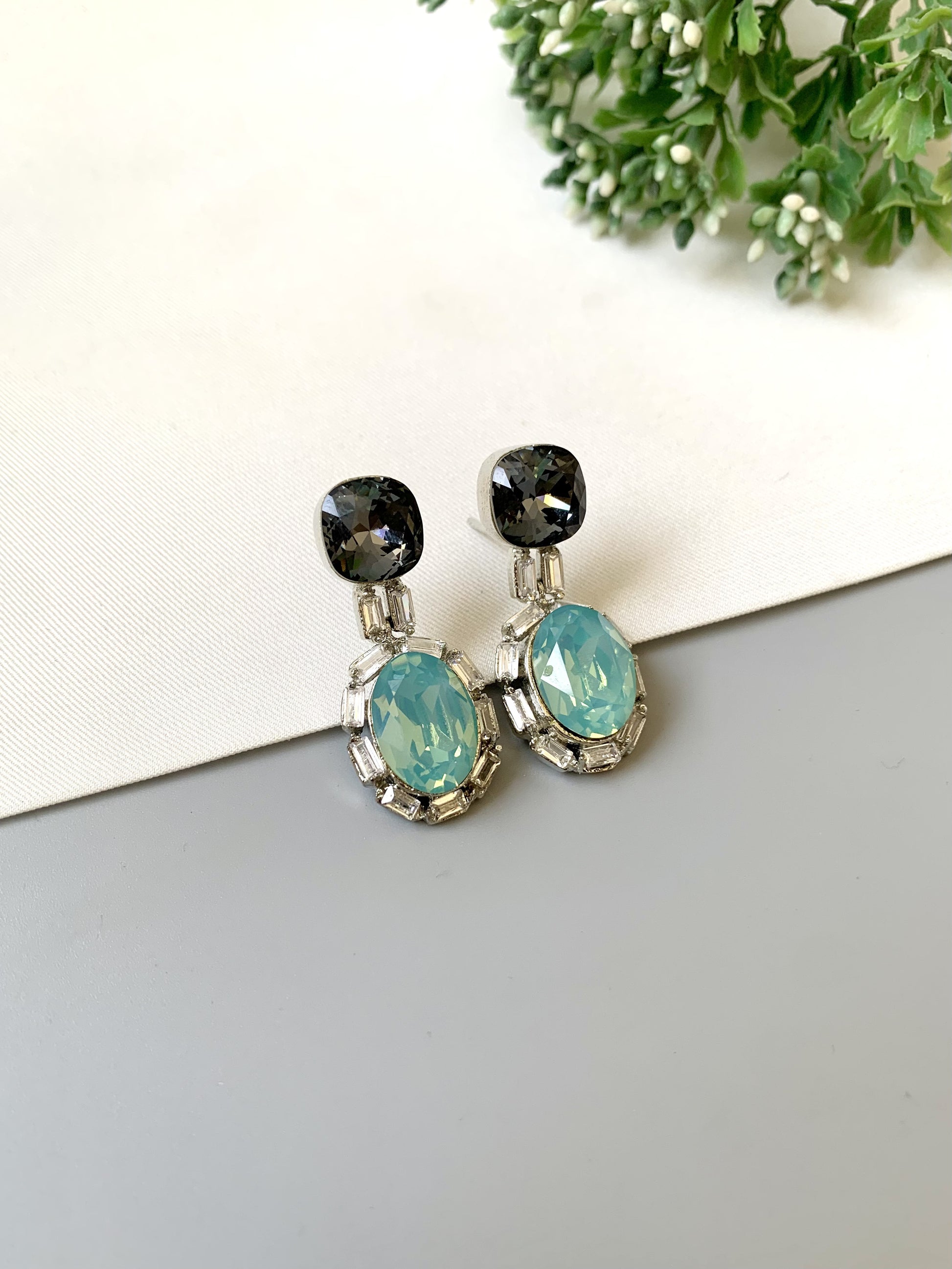 Pair of earrings with black and turquoise stones on a light gray surface with greenery in the background.
