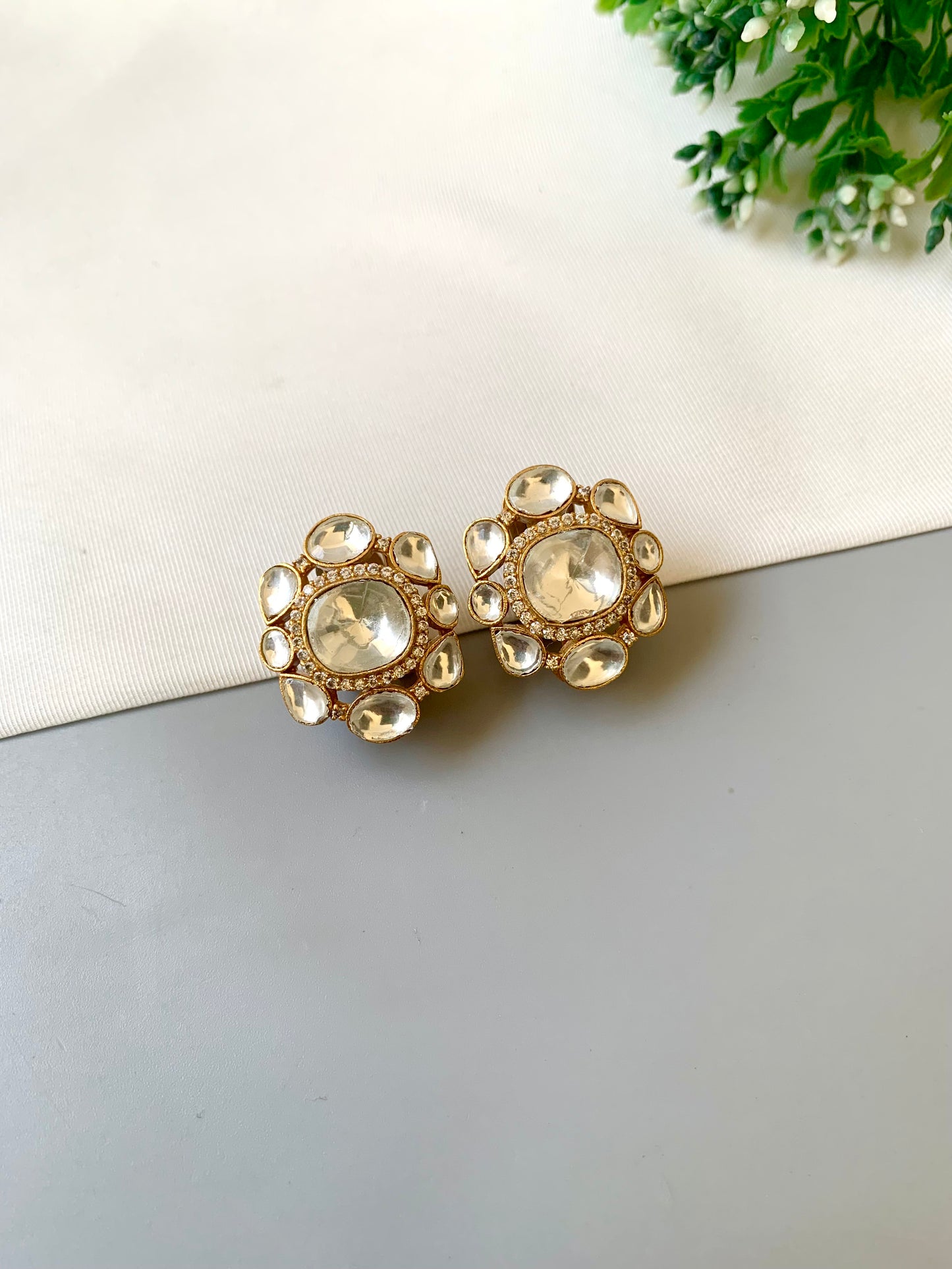 Gold earrings with clear gemstones on a light surface with greenery in the background