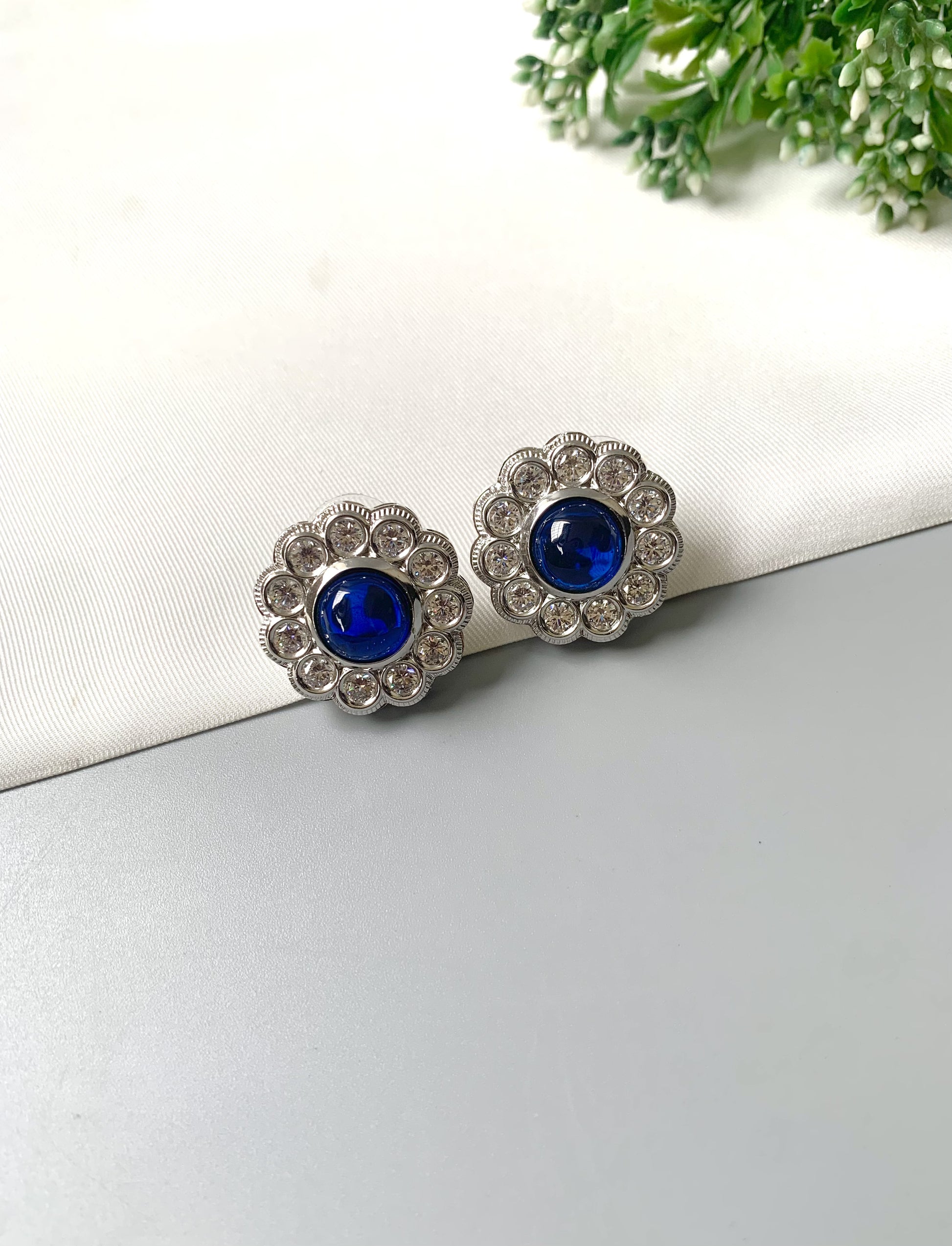 Pair of silver earrings with blue gemstones on a white surface with greenery in the background