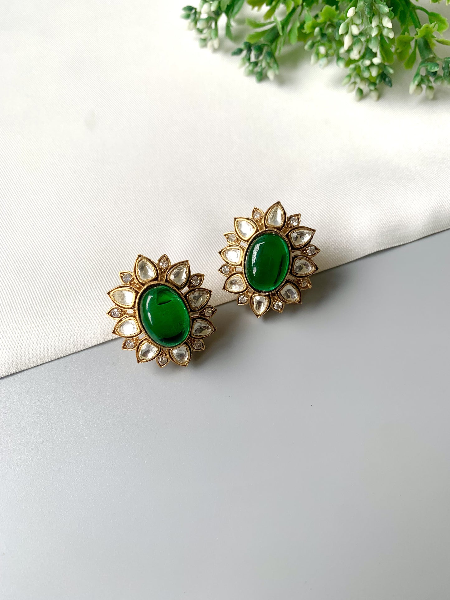 Pair of gold earrings with green gemstones on a white surface with green leaves in the background
