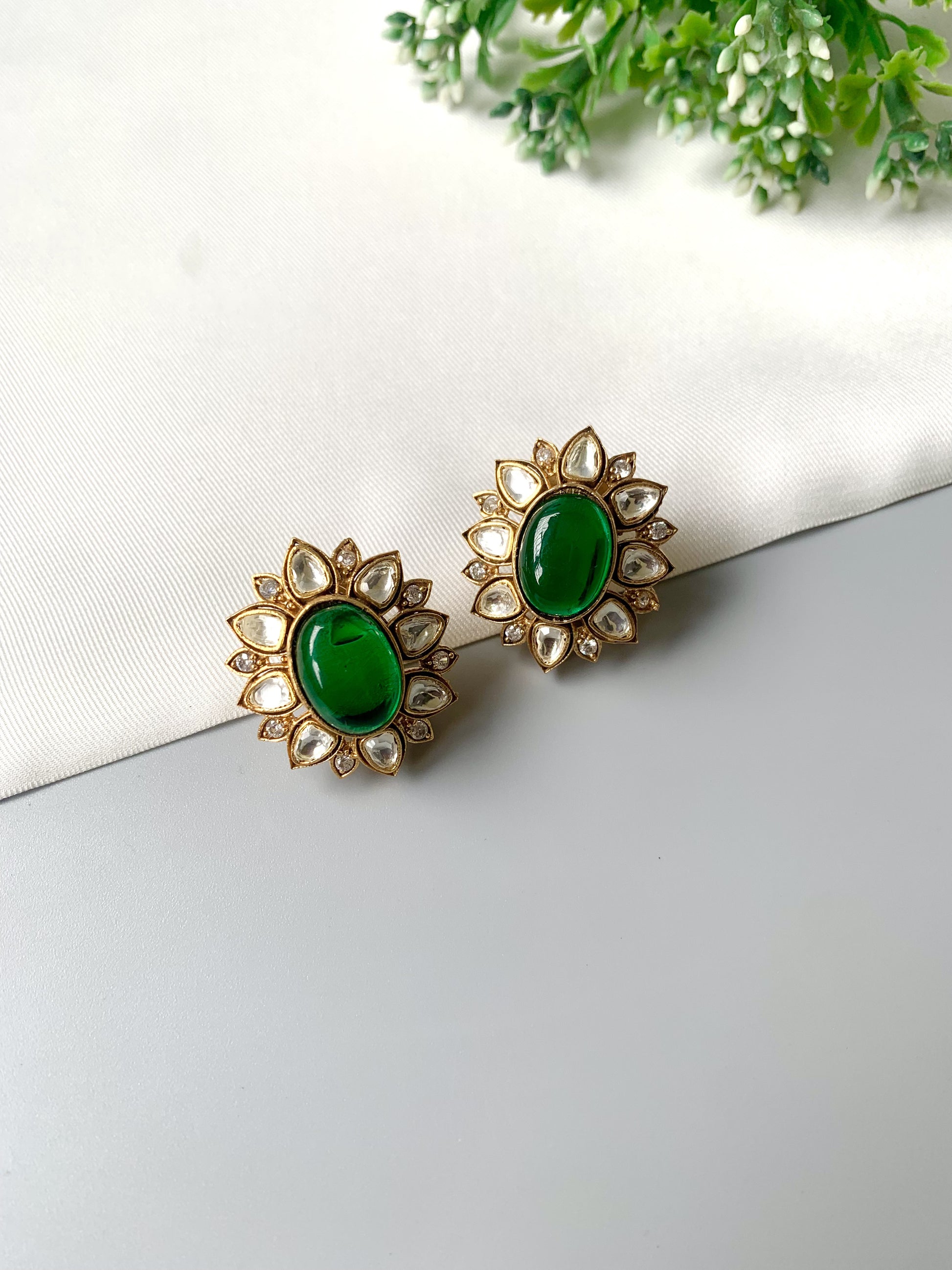 Pair of gold earrings with green gemstones on a white surface with green leaves in the background