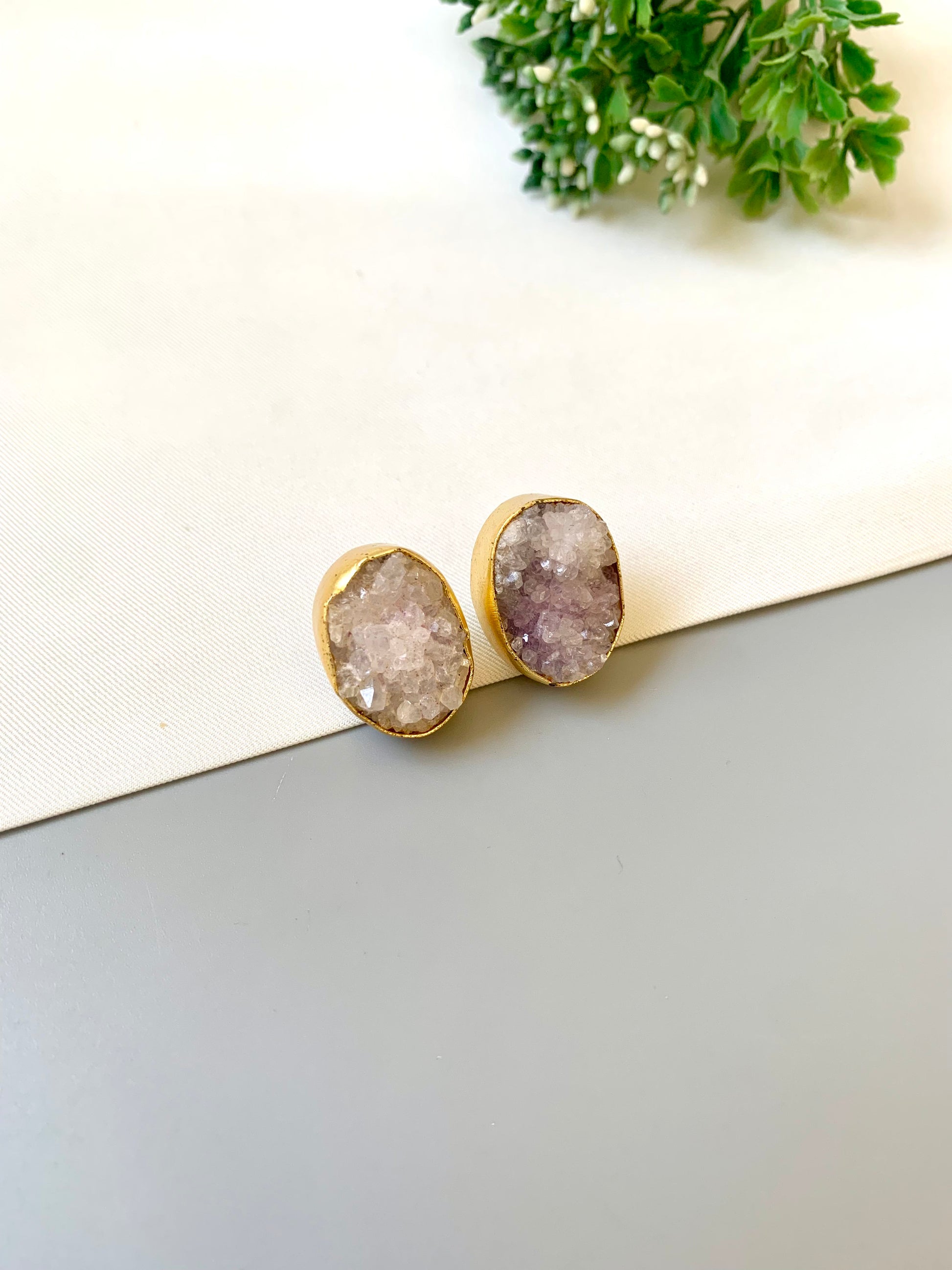 Gold-framed earrings with druzy stones on a light surface with greenery in the background