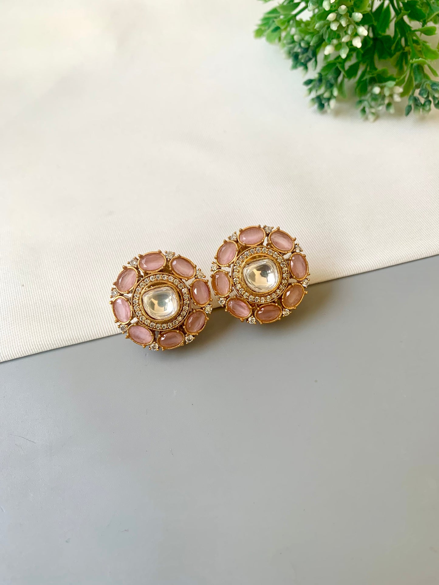 Pair of pink and gold earrings on a light gray surface with a white cloth and green plant in the background.