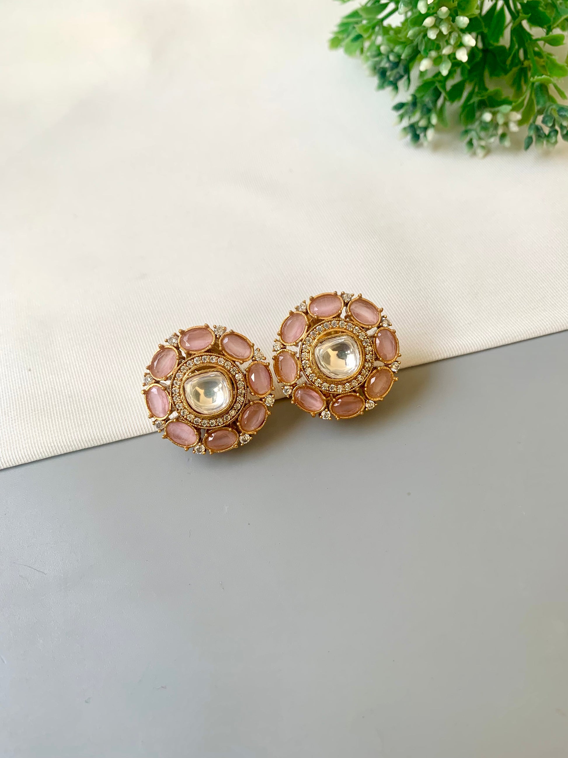 Pair of pink and gold earrings on a light gray surface with a white cloth and green plant in the background.