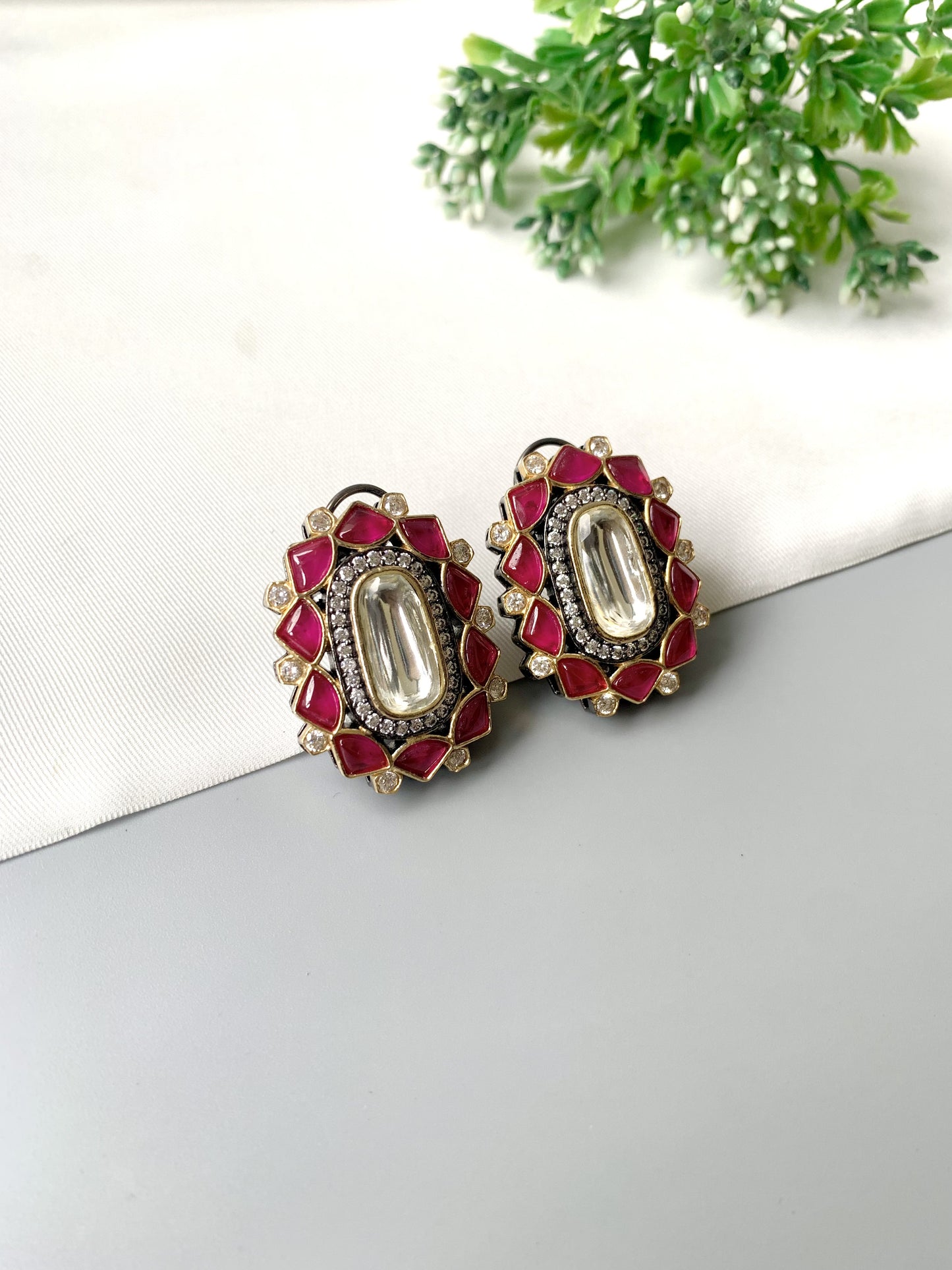 Decorative earrings with red and gold accents on a white surface with greenery in the background