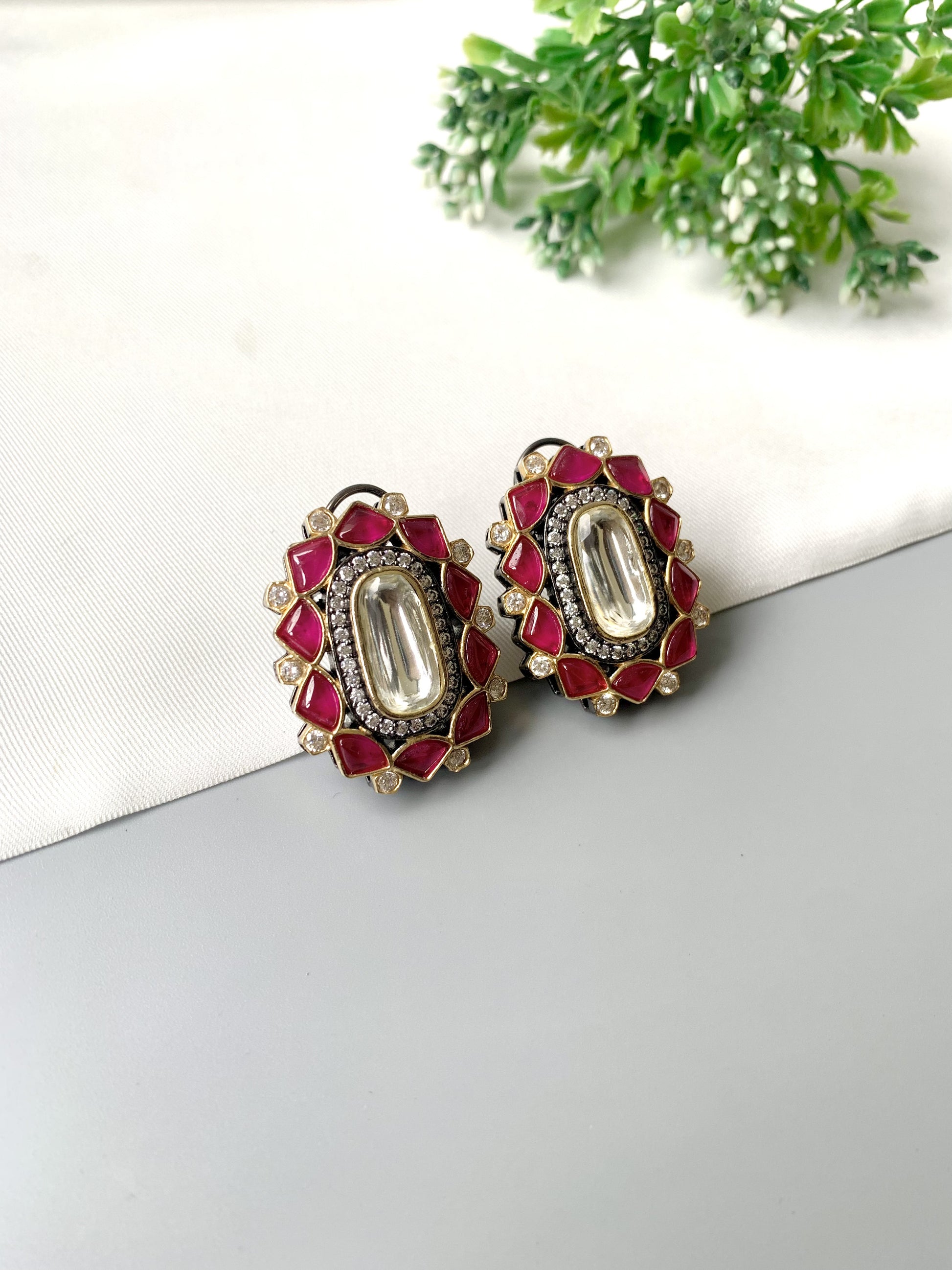 Decorative earrings with red and gold accents on a white surface with greenery in the background