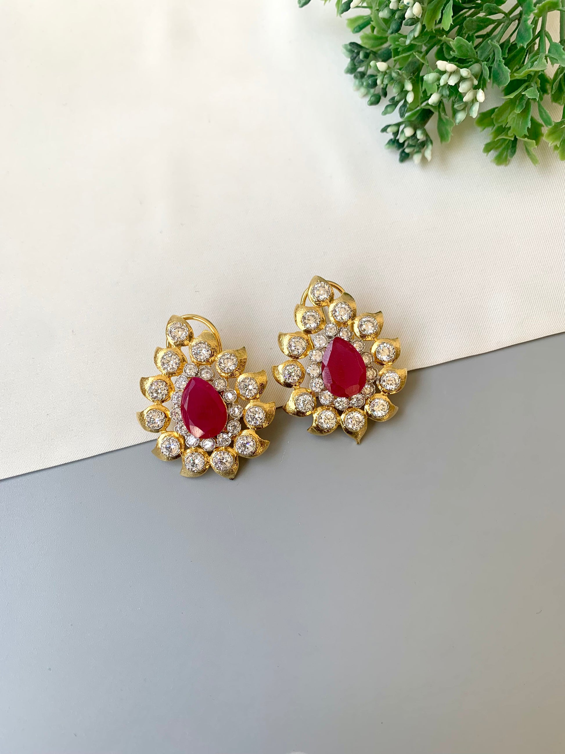 Gold earrings with red gemstones on a gray surface with greenery in the background