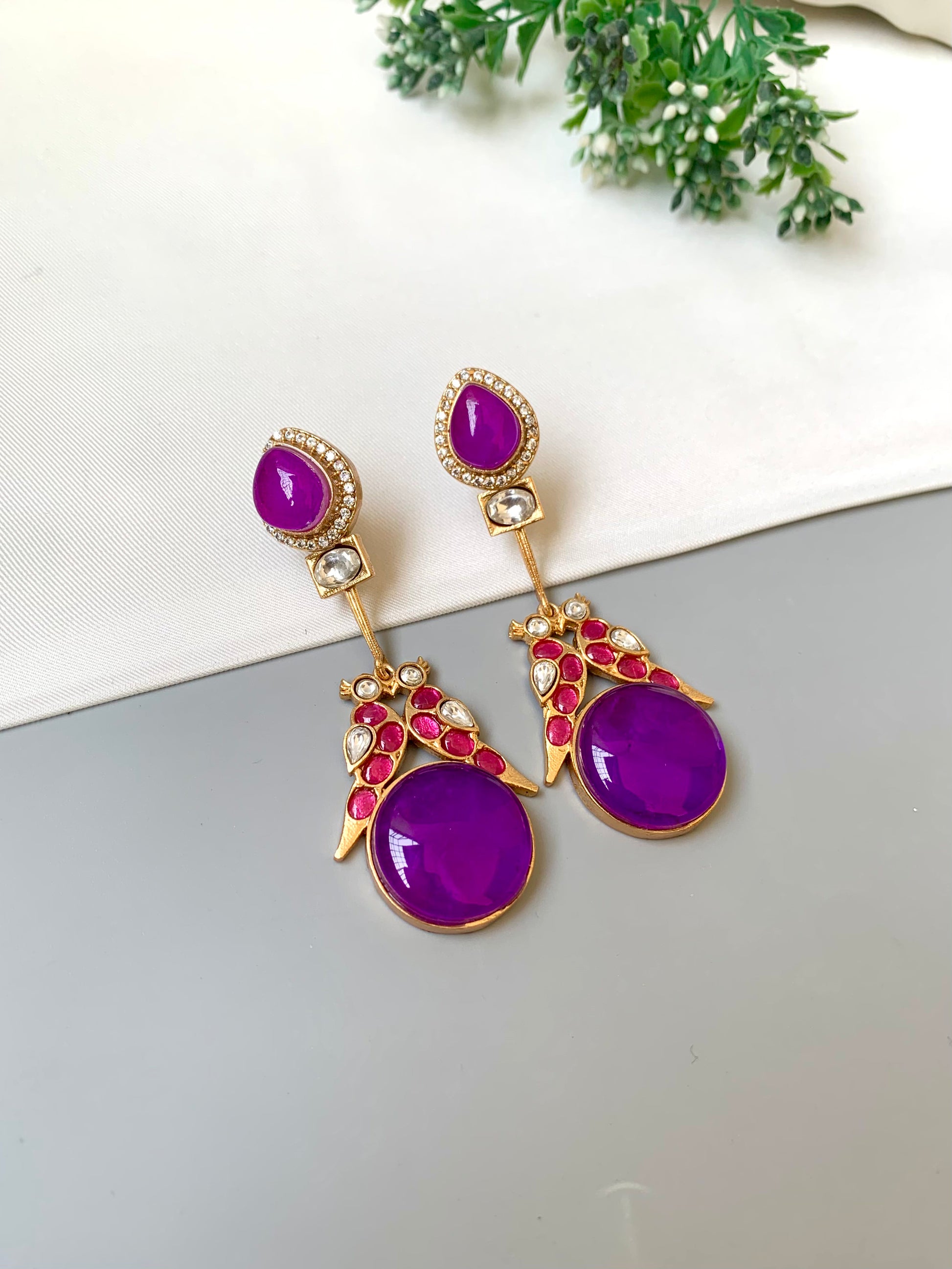 Purple earrings with gold and red accents on a light gray surface.