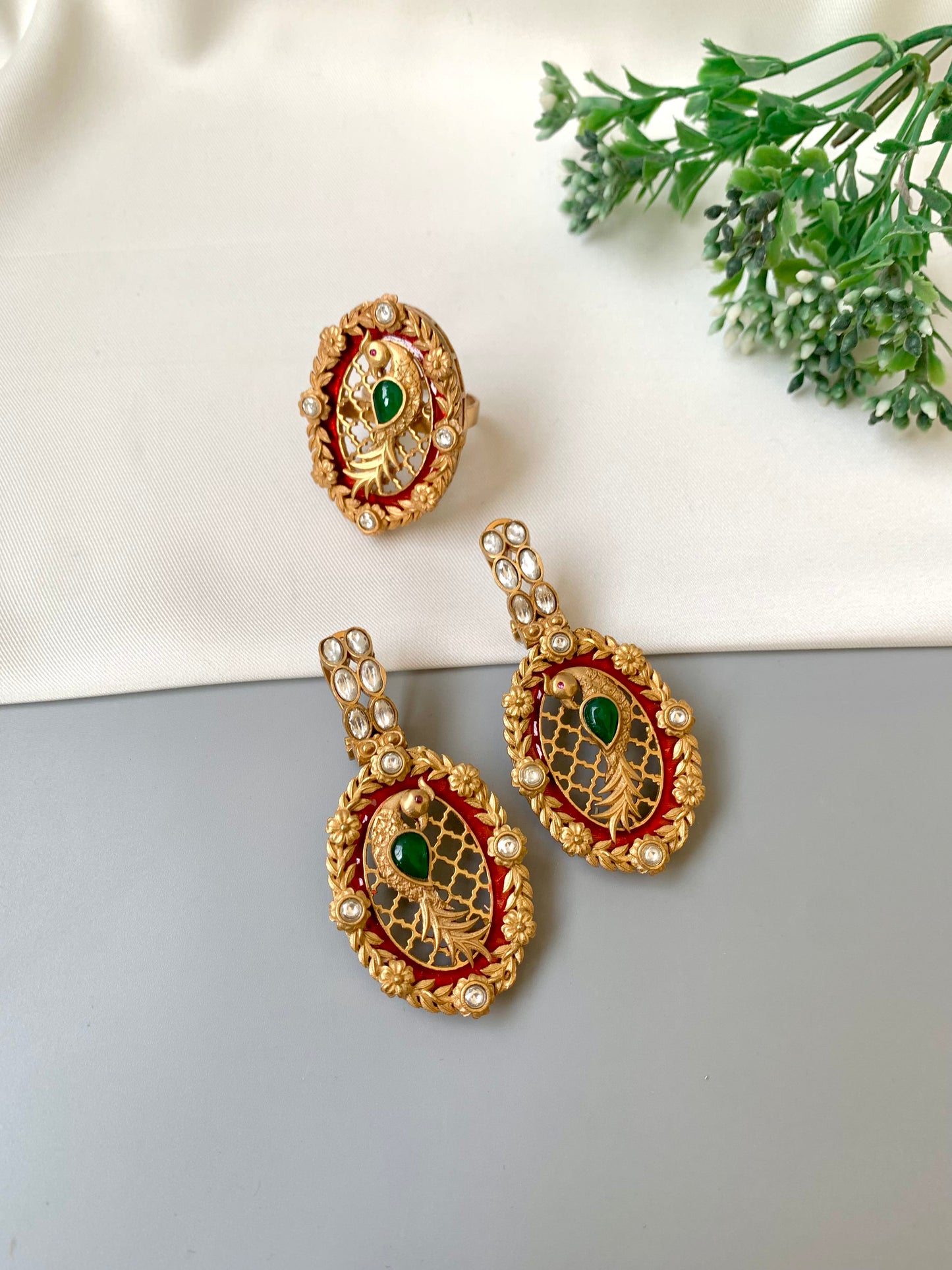 Ornate gold earrings with green and red accents on a light gray surface with a white cloth and green leaves in the background.