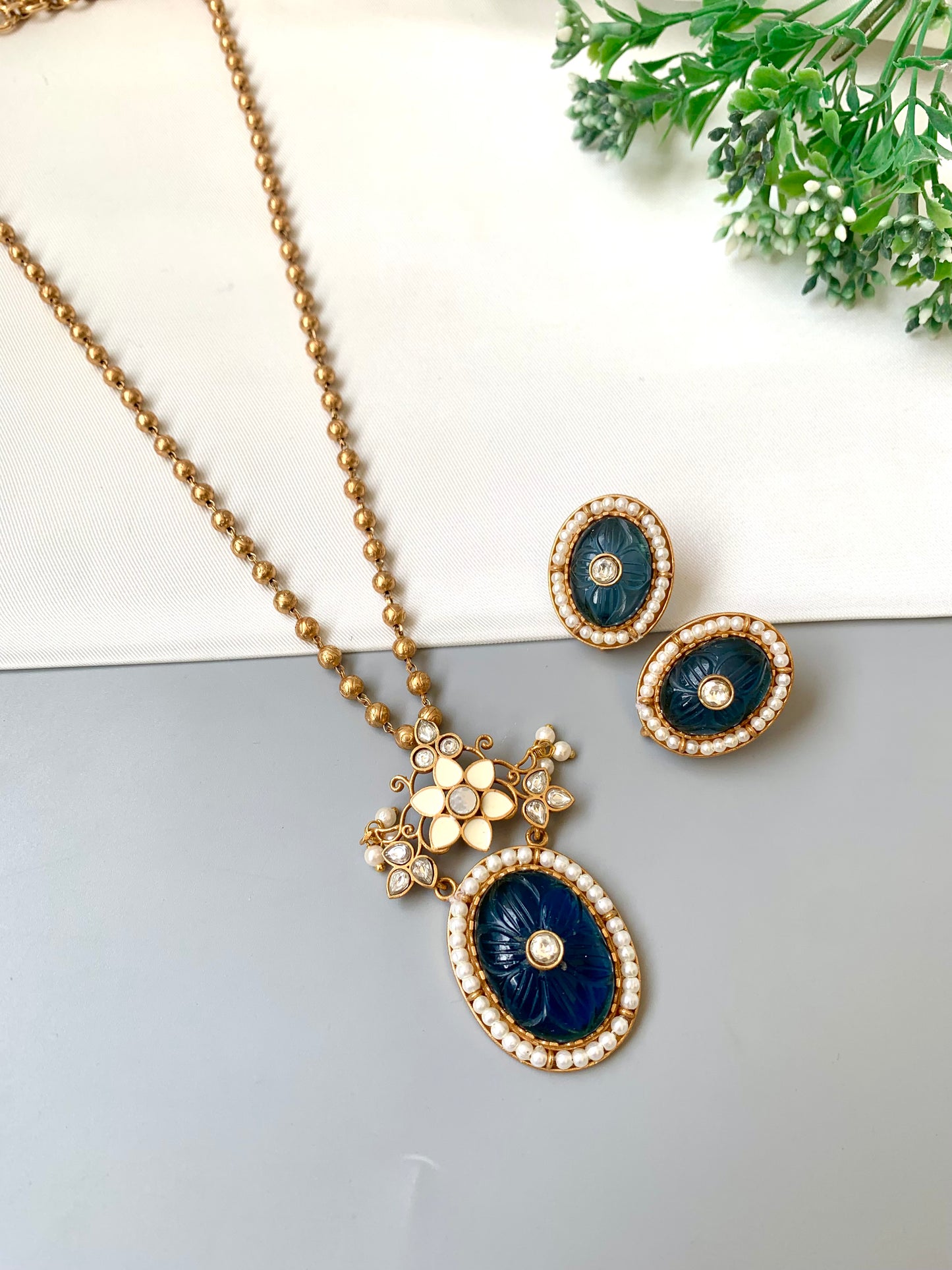 Navy blue and gold necklace and earrings on a light gray surface with greenery in the background