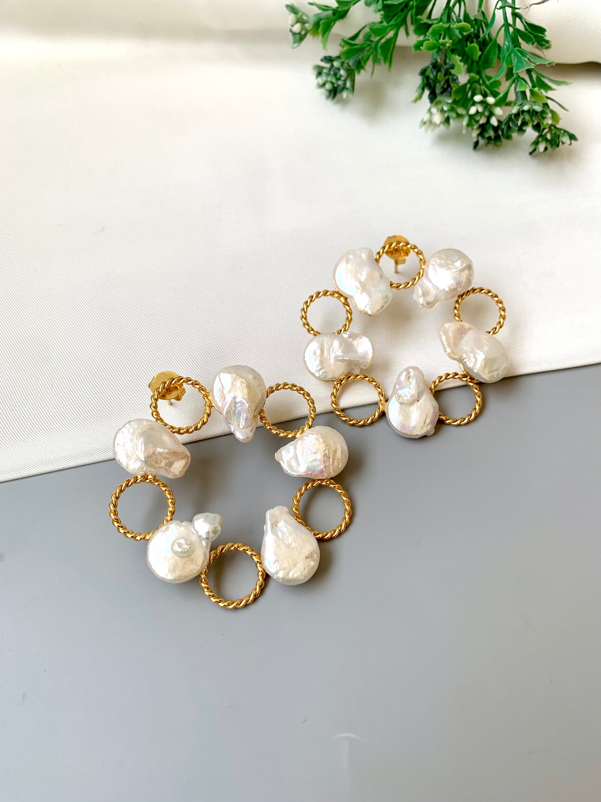 Pearl and gold hoop earrings on a light gray surface with greenery in the background